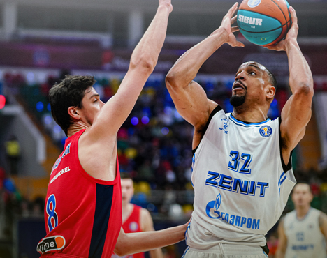 На изображении два баскетболиста: игрок в белой форме команды Зенит готовится выполнить бросок, против него защищается игрок в красной форме. Сцена происходит на баскетбольной площадке