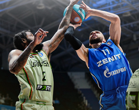 На изображении два баскетболиста борются за мяч. Один в форме синего цвета с надписью Зенит, другой в светлой форме с зелеными элементами. Они находятся в прыжке, вытянув руки вверх в спортивном зале